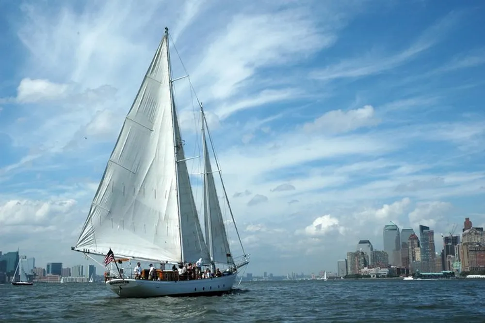 A sailboat is cruising on the water with a city skyline in the background under a partly cloudy sky