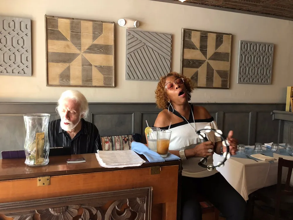 Two people sit at a restaurant table with decorative wooden art on the wall behind them the woman is capturing a playful moment with a facial expression and gesture while the man looks on slightly amused