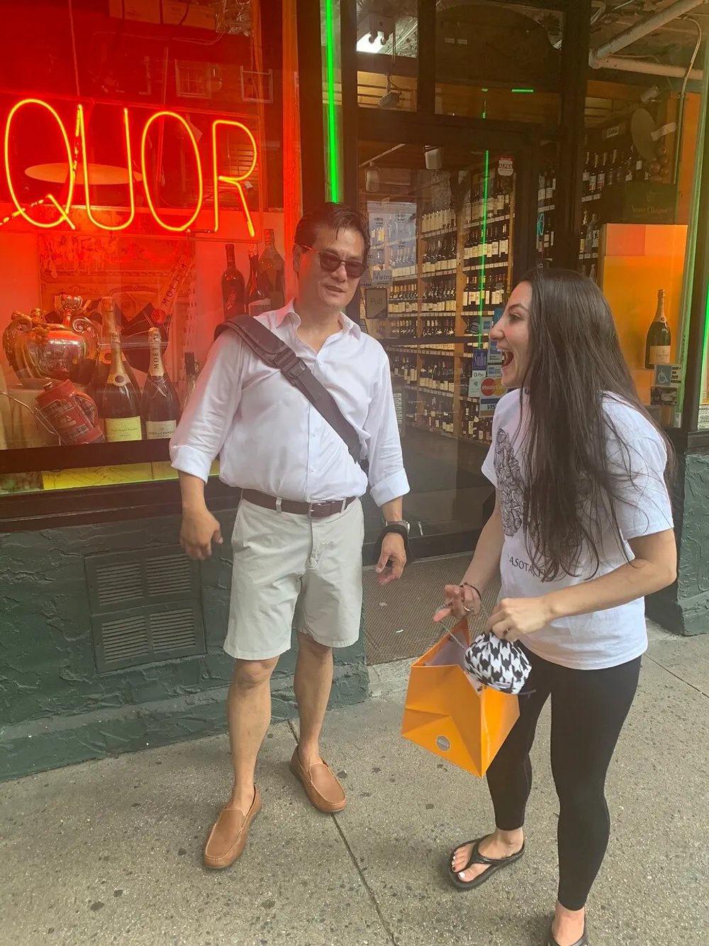 A man and a woman are standing in front of a store with a neon liquor sign sharing a moment of laughter or conversation