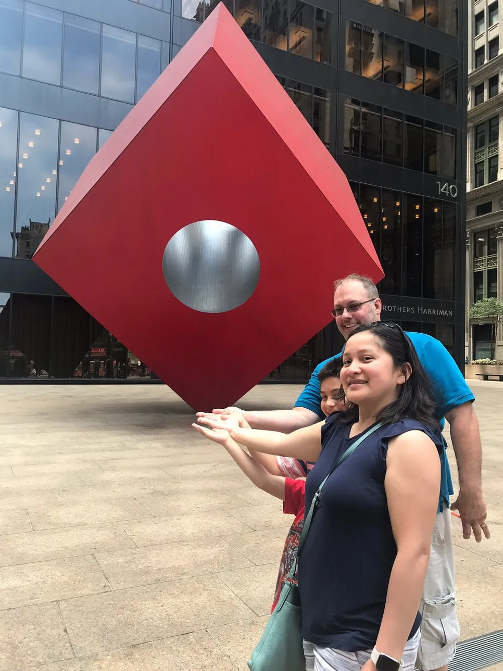Three people pose as if they are holding up a large red cube-shaped sculpture with a circular cut-out in an urban plaza