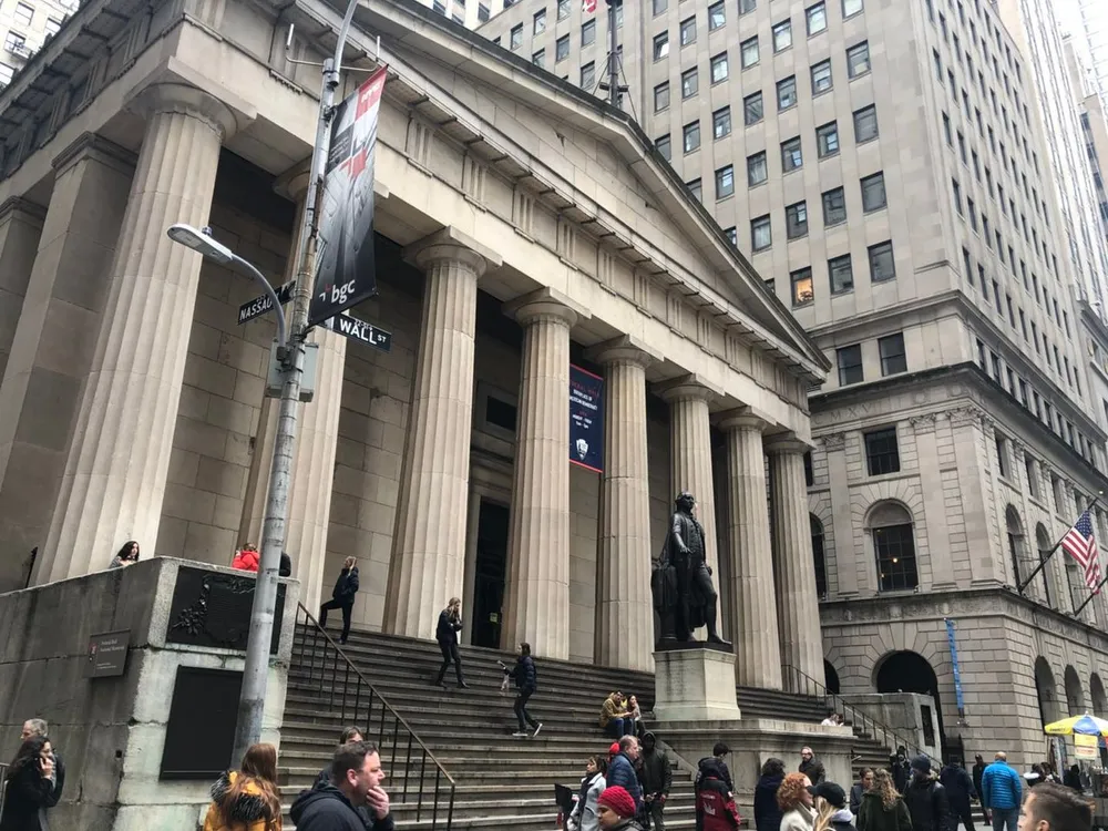 This image shows a bustling scene in front of a neoclassical building with large columns a statue and several people ascending and descending its front steps with street signs indicating the corner of Wall Street and Nassau Street