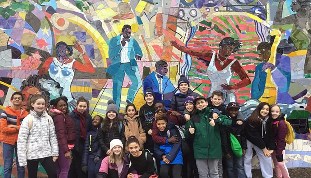A group of smiling children poses in front of a colorful mosaic wall featuring artistic depictions of people