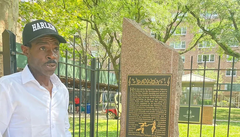 A man wearing a HARLEM cap stands near a historical marker detailing the significance of the legendary Savoy Ballroom in the development of jazz and popular dance cultures