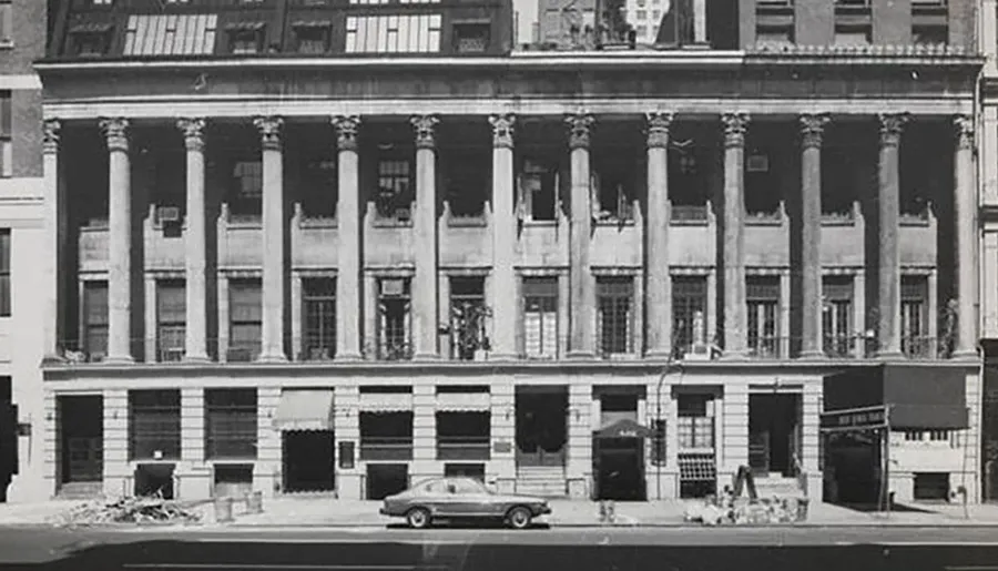 The image shows a black and white photograph of an older building with classical columns, several boarded-up windows, and a vintage car parked in front.