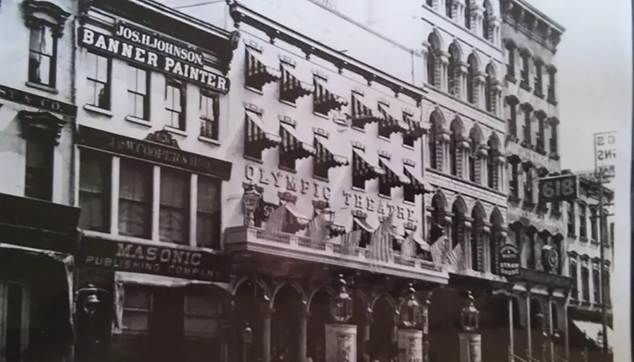 The image is a black and white photo of an old multi-story building with various business signs, including Jos. H. Johnson Banner Painter and Olympic Theatre, indicating a bustling commercial area from an earlier era.