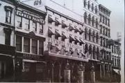 The image shows a historic black and white photograph of a city street with the facades of several buildings, one being the Olympic Theatre, surrounded by other businesses including a banner painter and a Masonic publishing company.