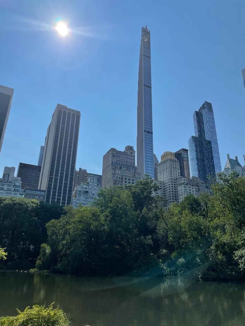 The image shows a view of a tranquil body of water surrounded by lush greenery in the foreground with towering skyscrapers against a clear blue sky in the background dominated by a particularly tall and slender building