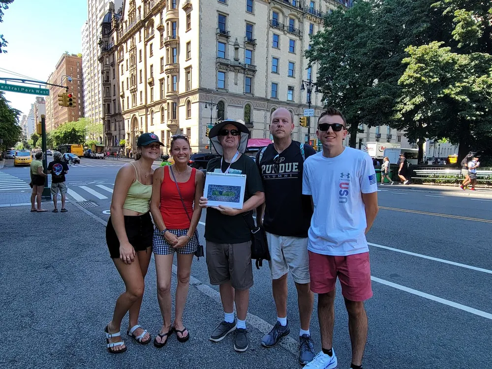 A group of five people is smiling for a photo on a sunny day at a city crosswalk with trees and buildings in the background