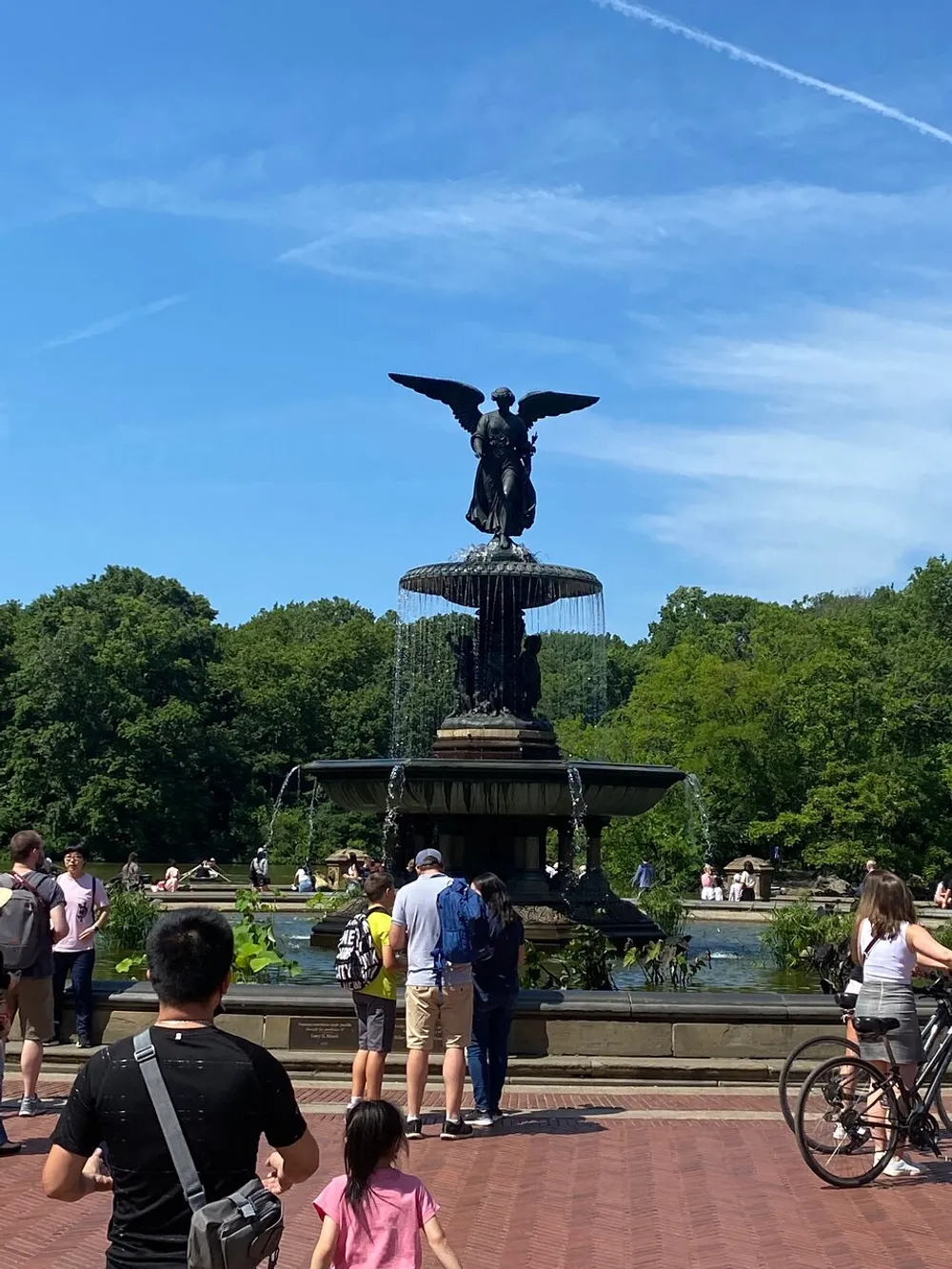 Tourists and locals alike enjoy a sunny day around a large ornate fountain with an angel statue at the center likely in a public park