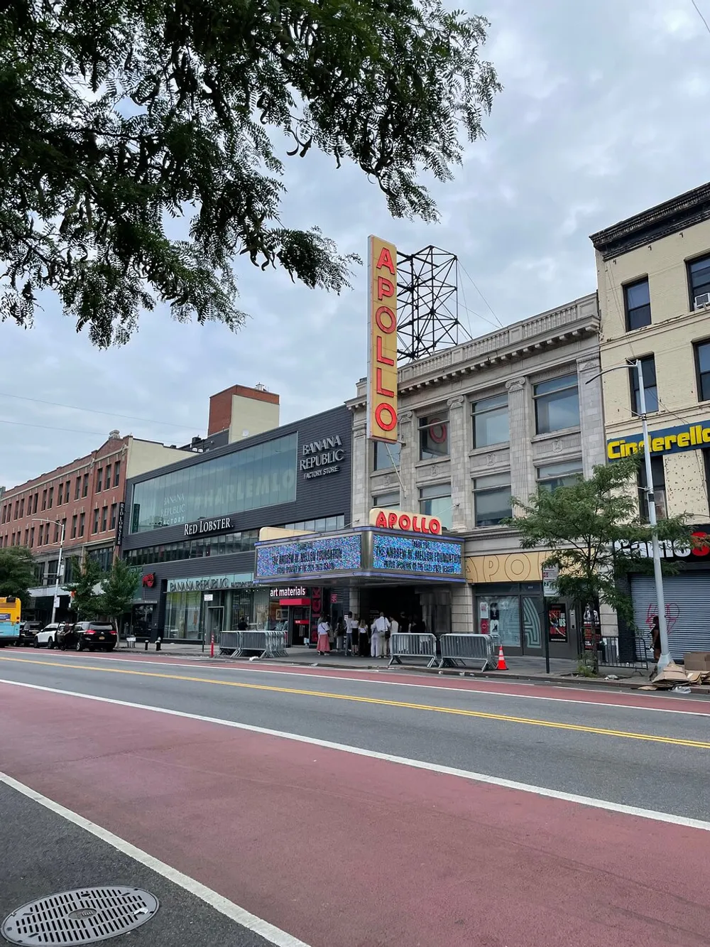 The image shows the iconic Apollo Theater with its notable neon sign located on a city street with pedestrians and various storefronts under an overcast sky