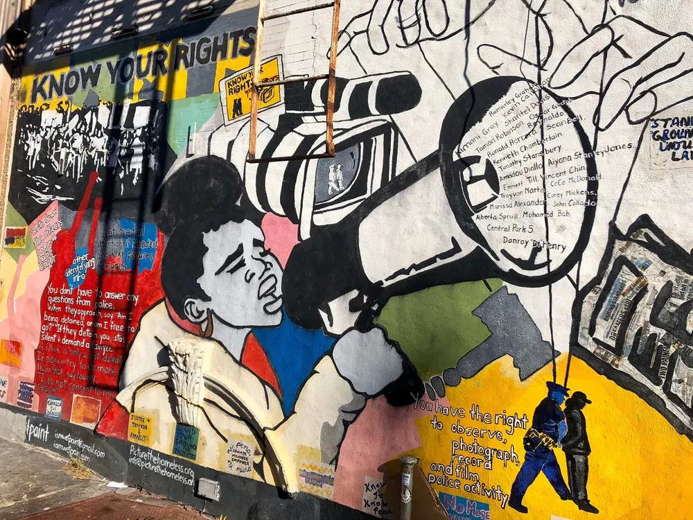 The image shows a colorful mural with messages about civil rights and empowerment featuring a figure with a megaphone and names within an outstretched hand urging people to know their rights