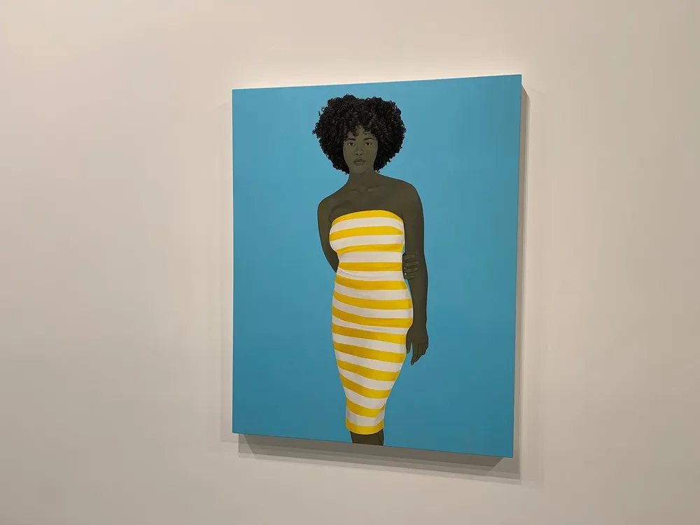 The image shows a painting of a woman with dark curly hair wearing a yellow and white striped dress set against a light blue background framed and displayed on a white wall