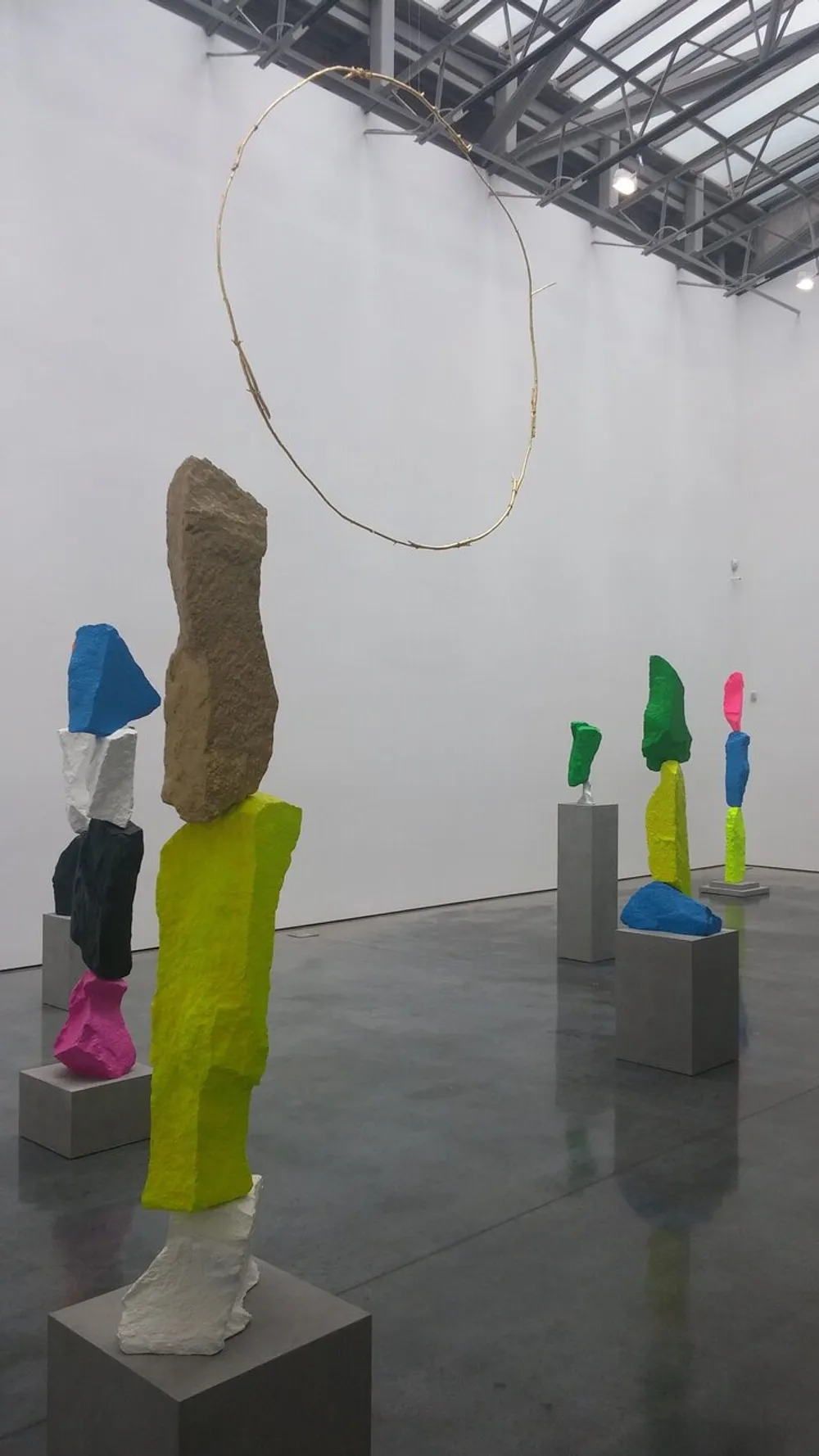 The image shows a contemporary art installation featuring colorful abstract sculptures on pedestals with a large golden loop hanging from the ceiling in the background
