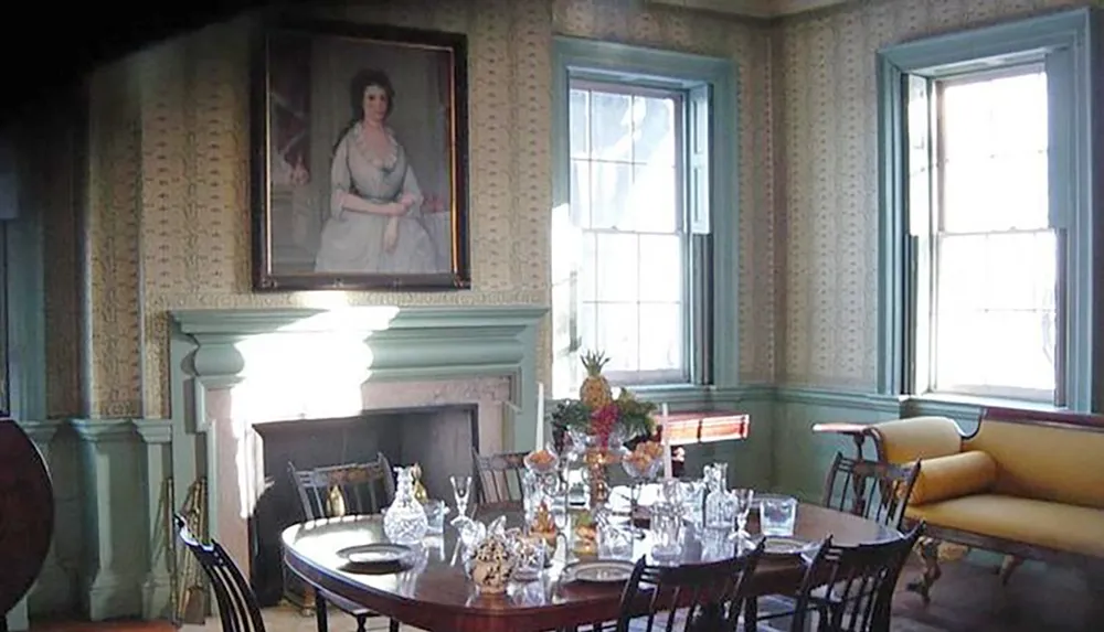 The image shows an elegantly set historical dining room with a large oval table glassware a fireplace traditional furniture a portrait painting on the wall and two windows allowing in natural light