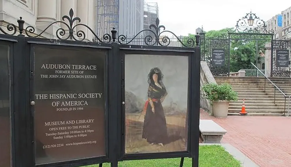 The image shows a gated entrance to a cultural institution with informational signage about The Hispanic Society of America and a portrait on display