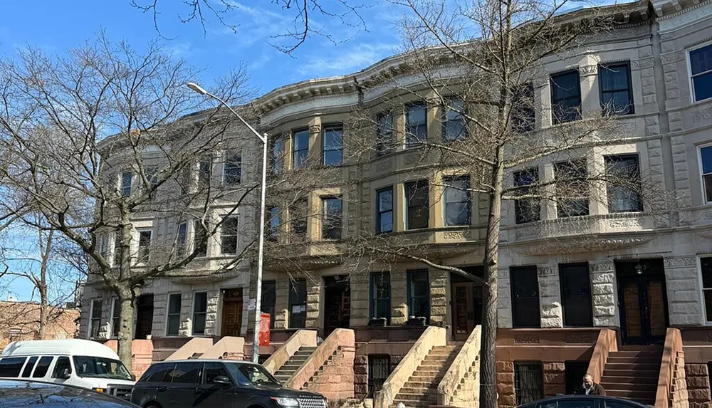 The image shows a row of brownstone houses with staircases to the entrance on a sunny day with leafless trees and parked cars in the foreground