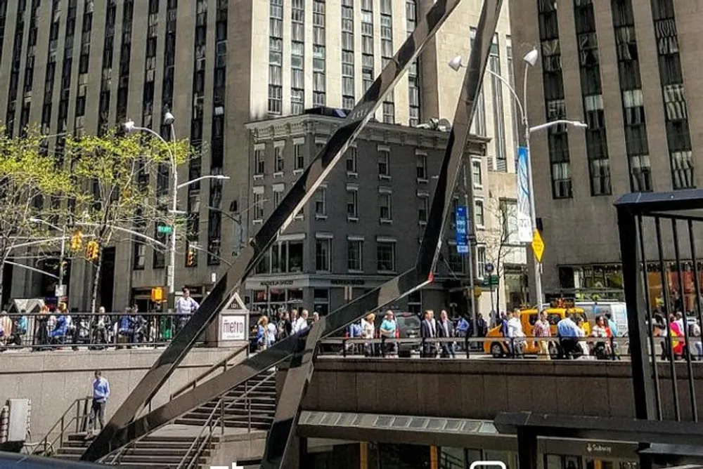 This image captures a bustling city scene with a crowd of people on a sidewalk modern buildings a subway entrance and a distinctive sculptural metallic structure under a clear blue sky