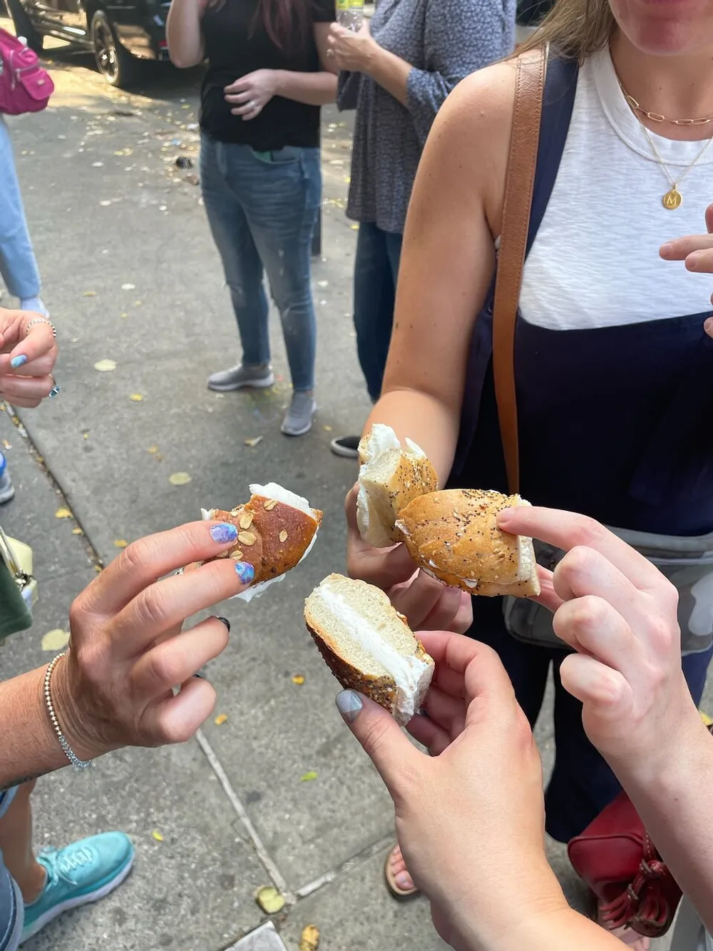 A group of people are holding and presumably sharing different varieties of bagel sandwiches outdoors