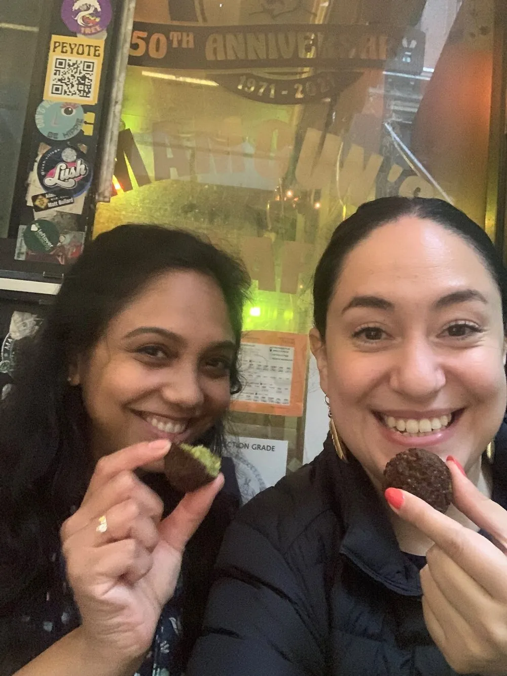 Two individuals are smiling at the camera holding up what appears to be small treats with a window reflecting a neon sign and various stickers in the background