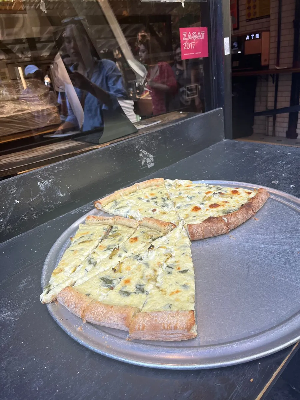 Three slices of white pizza with a variety of toppings sit on a metal pizza tray on a dark countertop with reflections of people visible in the glass window of a pizzeria behind it