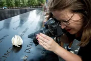 A woman is photographing a white rose placed on a name at a memorial with engraved names, capturing a poignant moment of remembrance.