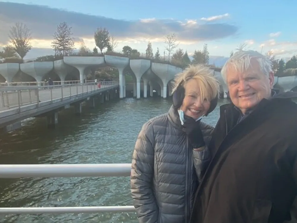 A smiling couple poses for a photo in front of a modern bridge over a body of water with a cloudy sky above them