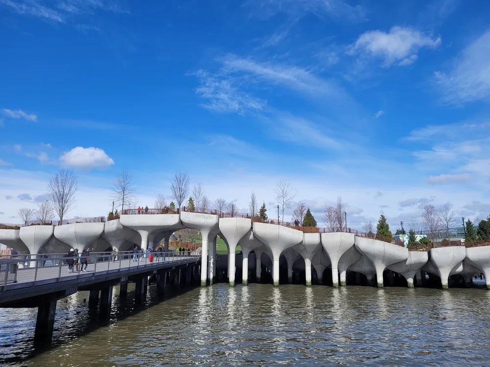 A unique bridge with an organic sculptural design spans a body of water under a blue sky with scattered clouds