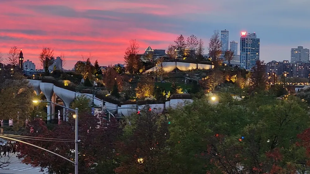 The image displays a vibrant sunset with shades of pink and orange in the sky illuminating an urban park with modern architecture and the surrounding cityscape in the background