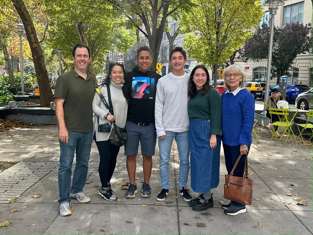 A group of six people are smiling for a photo on a sunny day with trees and a city street in the background