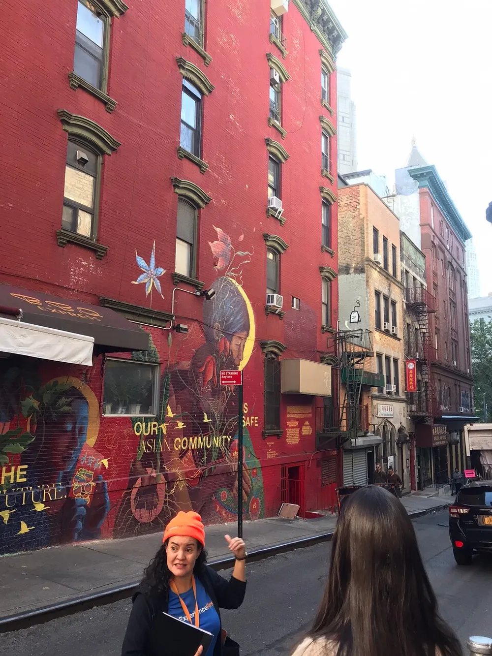 The image shows a vibrant street mural with the message Our Asian Community is Safe on a red building with a tour guide in an orange hat gesturing towards the artwork while speaking to her audience