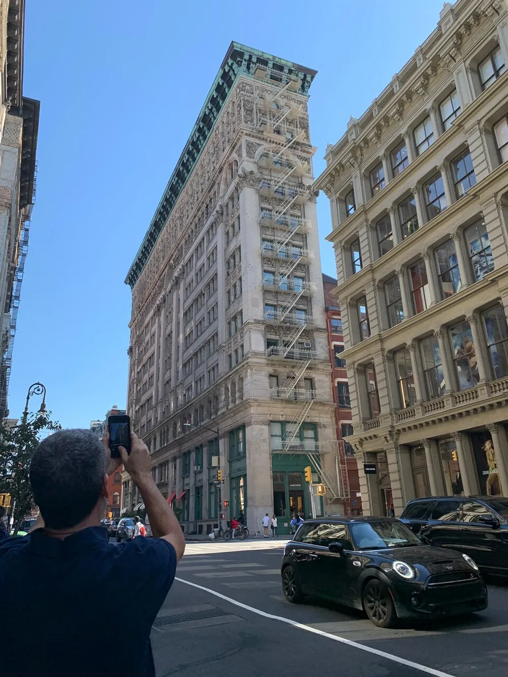 A person is taking a photo of a distinctive flatiron-style building on a sunny day in a bustling city street