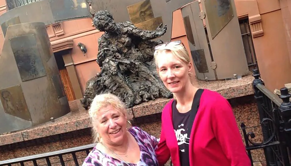 Two women are smiling for a photo beside a bronze statue on a city street