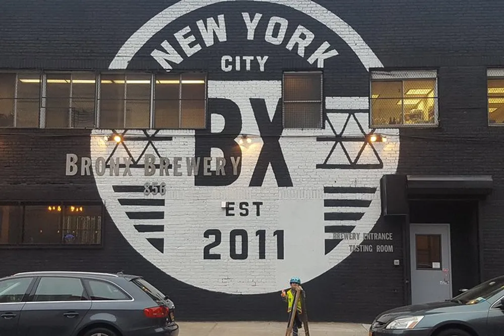 The image shows the exterior of a building painted black with a large white circular logo that reads New York City BX Bronx Brewery EST 2011 indicating the establishment is a brewery in the Bronx New York City established in 2011 with a person wearing a helmet standing in front of it