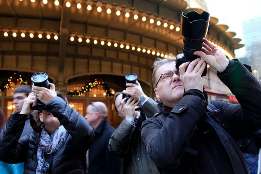 A group of people are looking up and taking photographs with various cameras against a backdrop of festive lights