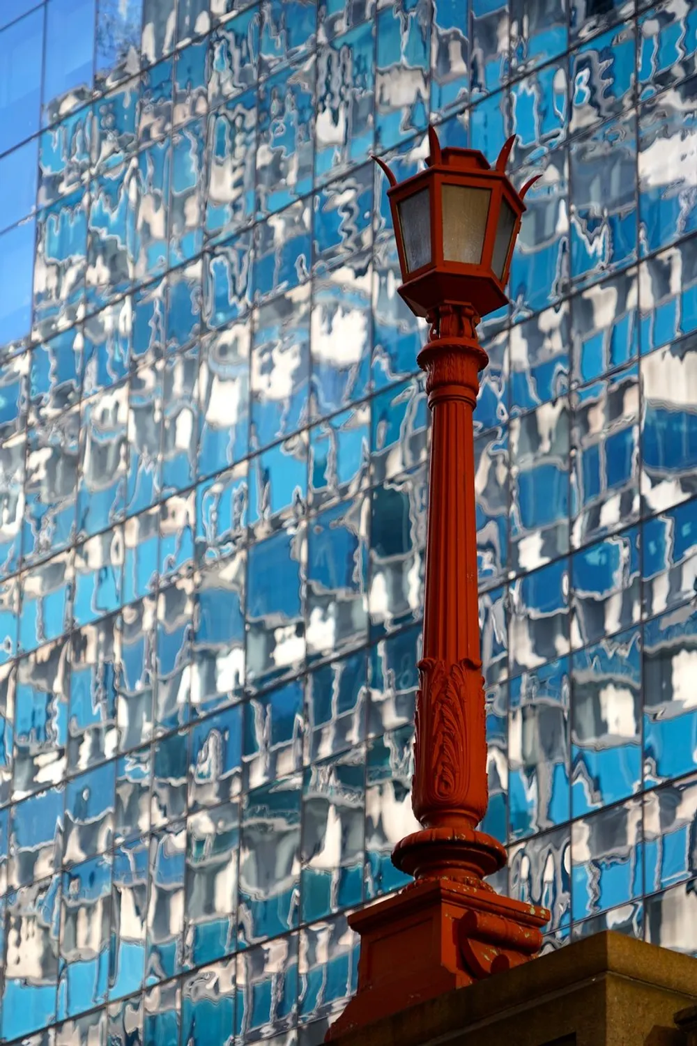 A red street lamp stands in the foreground with a distorted reflection of the blue sky on a glass building behind it