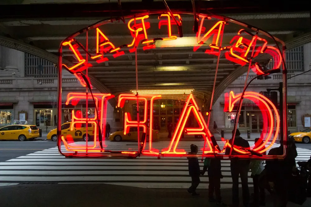 This image captures a vibrant red neon sign that reads ASTOR PLAZA in reverse due to the perspective with silhouettes of people a crosswalk and yellow taxis in the background suggesting an urban setting likely at dusk
