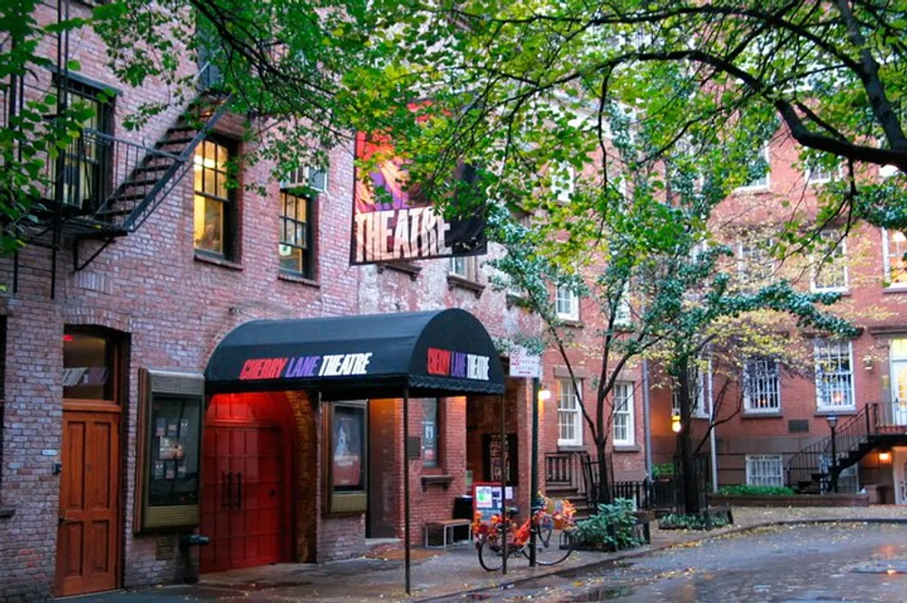 The image shows the Cherry Lane Theatre with its classic red brick facade and black awnings tucked away on a leafy street that gives off a quaint and historic atmosphere