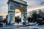 The image features the iconic Washington Square Arch in New York City with people crossing the street and walking around the park at twilight.