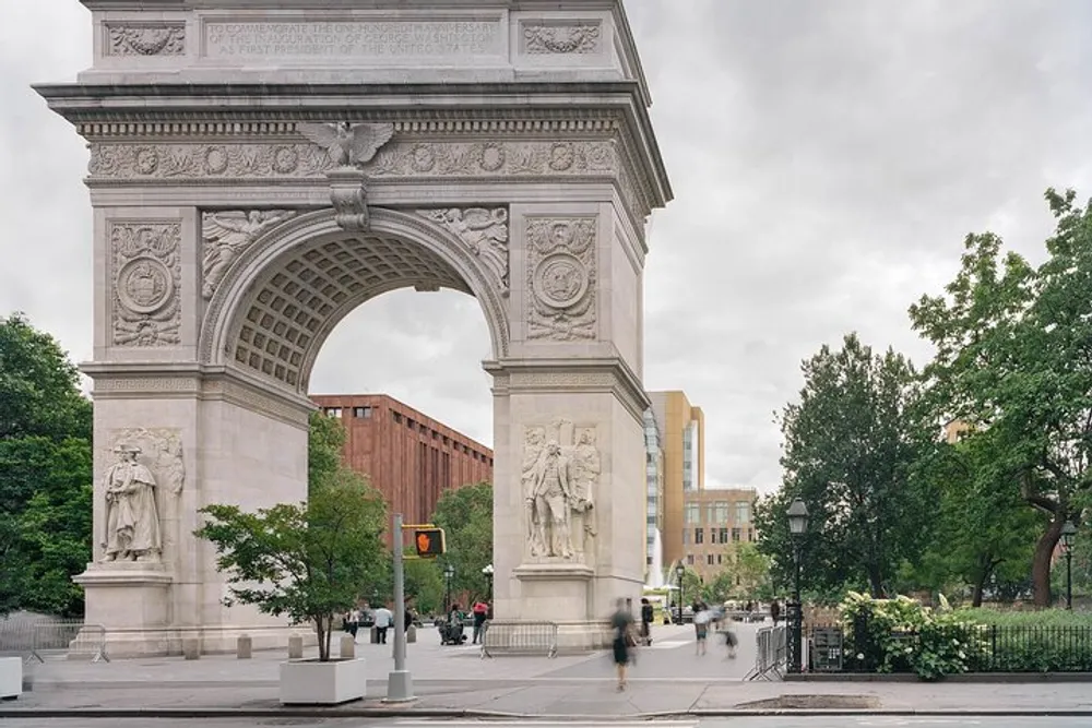 The image shows the Washington Square Arch an iconic marble triumphal arch in Washington Square Park New York City with trees pedestrians and buildings in the background