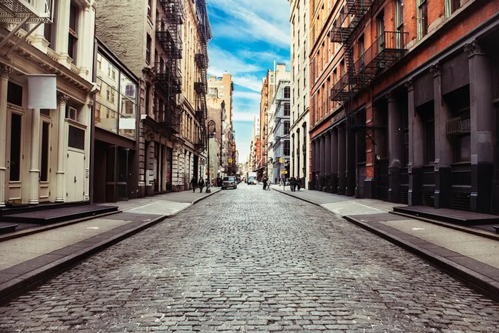 The image shows a classic cobblestone street flanked by historic buildings under a partly cloudy sky likely situated in an old urban neighborhood