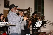 A man in a hat plays the trombone with a band of brass musicians in the background.