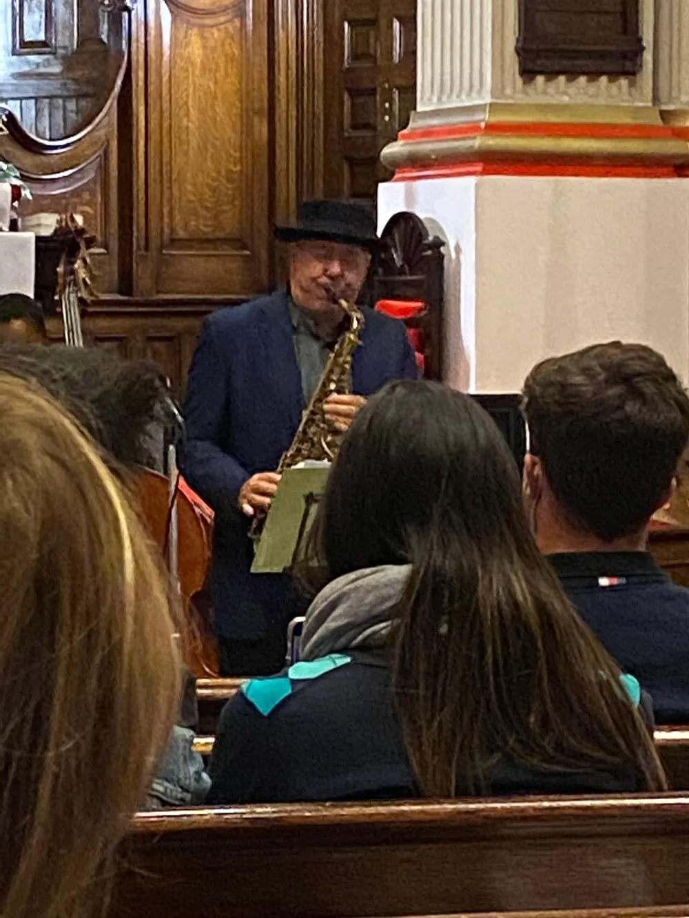 A man wearing a hat is playing the saxophone in front of an audience seated in wooden pews