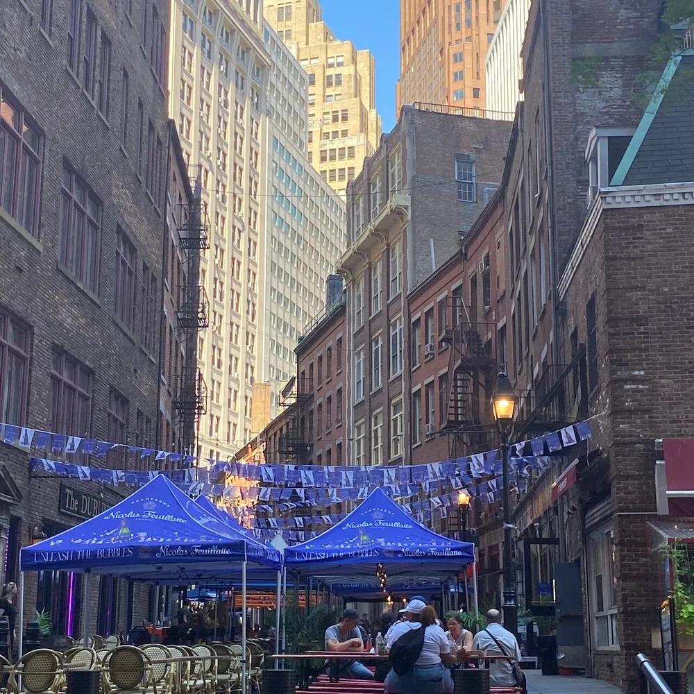 This image captures a vibrant outdoor dining scene on a narrow city street flanked by tall buildings and adorned with blue umbrellas and bunting