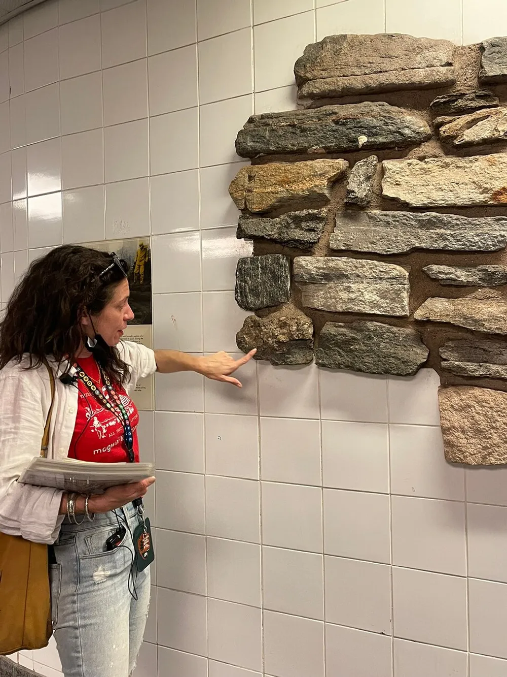 A woman is pointing to a section of a wall that transitions from smooth white tiles to a rough stone surface giving the illusion that the stones are emerging from the tiled wall