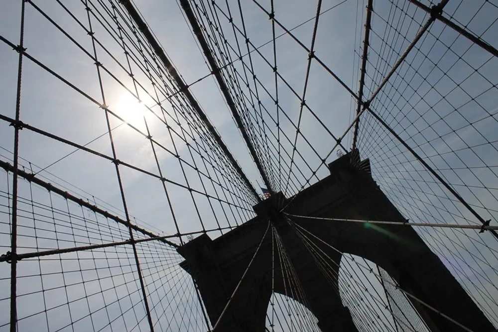The image captures a striking view of the sun peeking through the network of cables of a suspension bridge against a clear sky