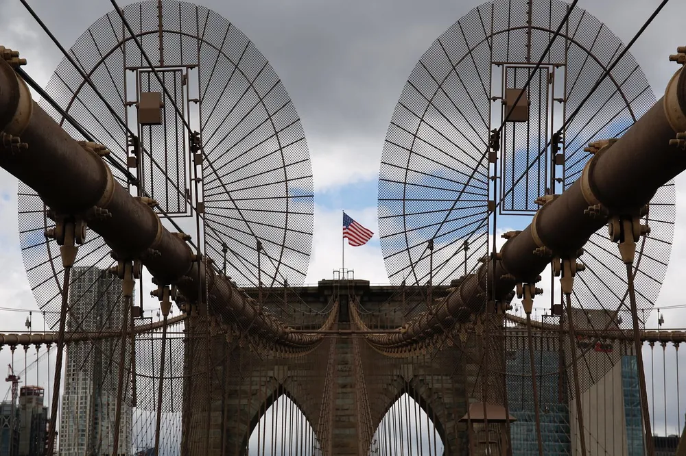 The image showcases a symmetrical view of the iconic Brooklyn Bridges stone arch and cable structure with an American flag proudly flying in the center against a cloudy sky