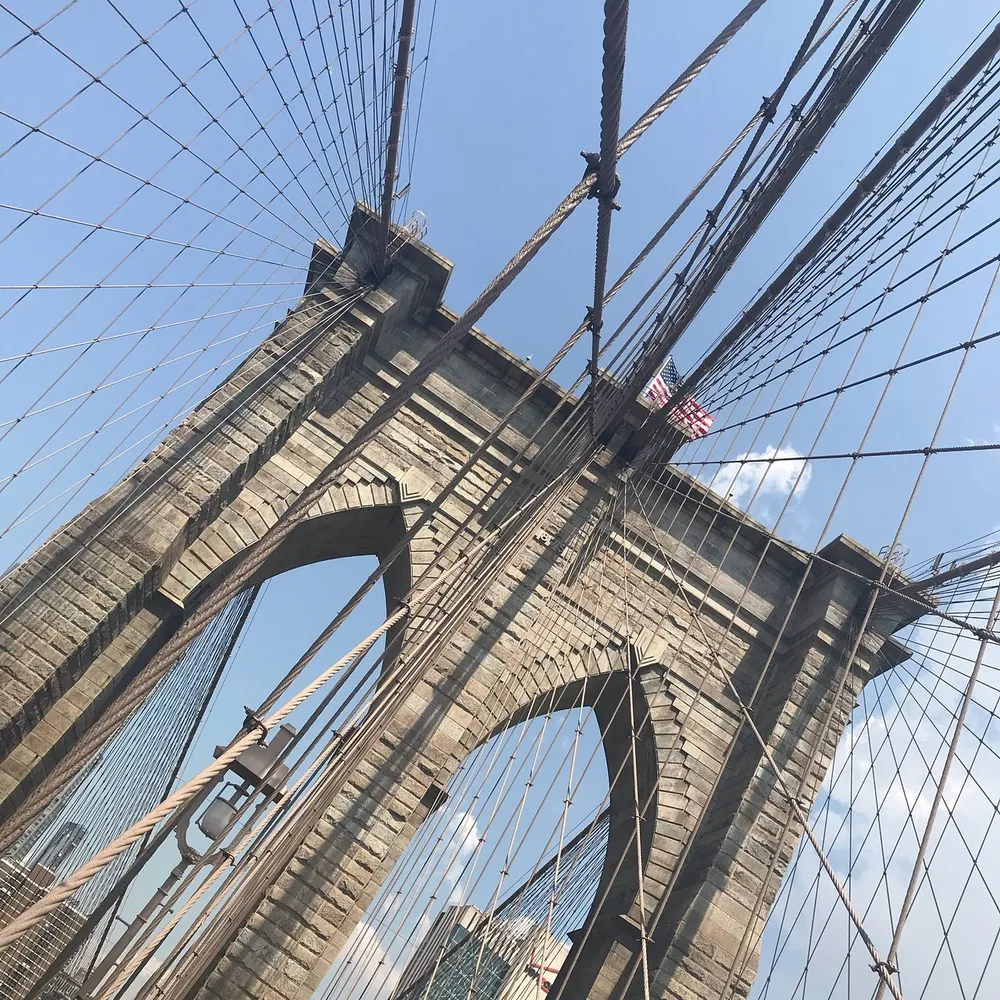 The image shows a perspective view of the Brooklyn Bridge with its distinctive cables and stone arches against a clear sky