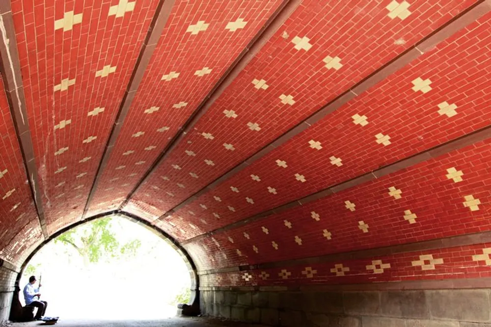 A person is sitting under a curved red-patterned brick underpass or tunnel with daylight showing at the end