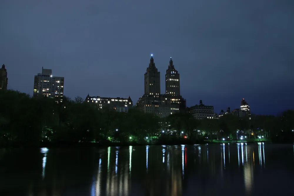 The image depicts a serene nighttime view of illuminated buildings reflecting on the calm water likely captured from within a park