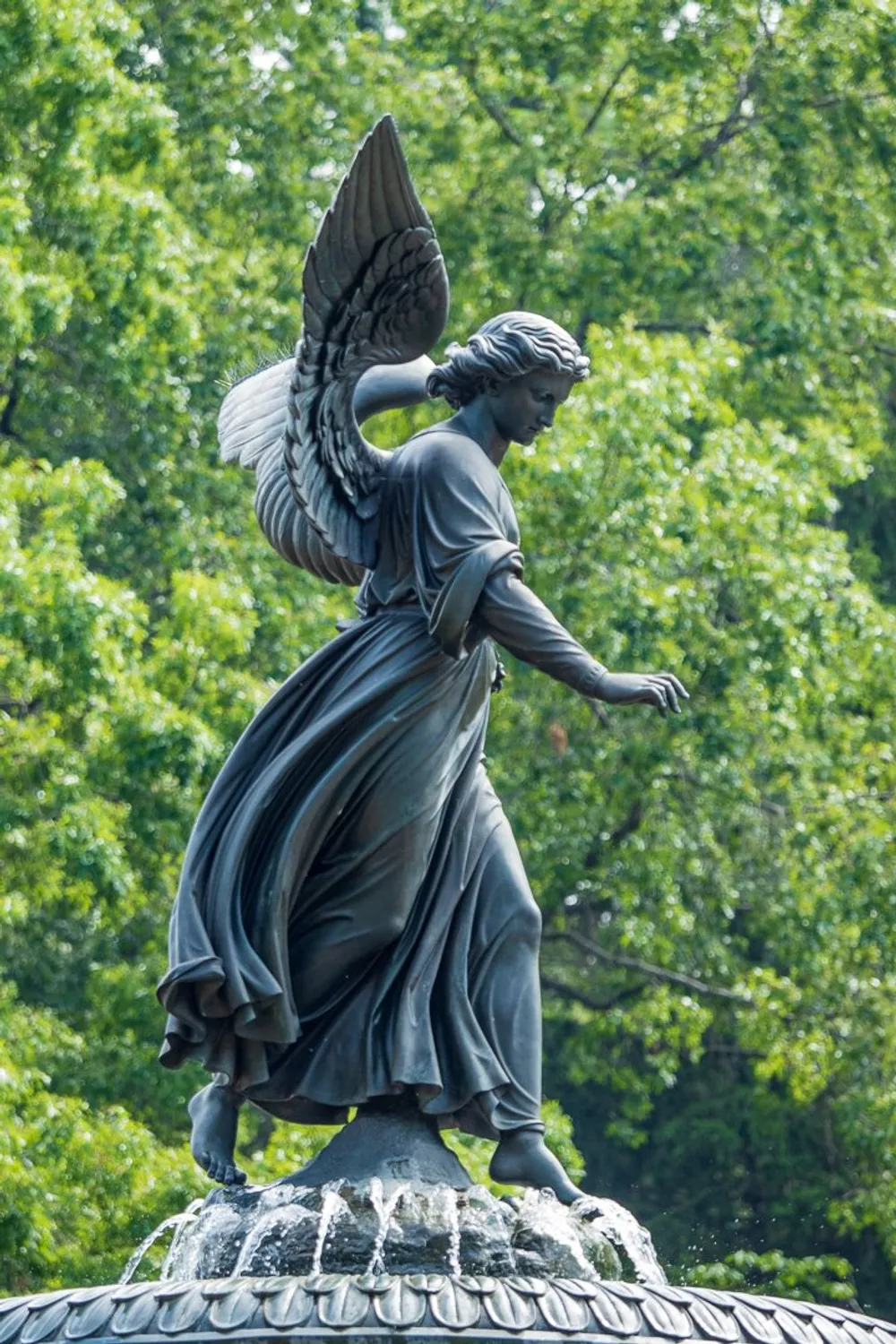 The image shows a bronze statue of an angel with wings standing on top of a fountain against a backdrop of green foliage